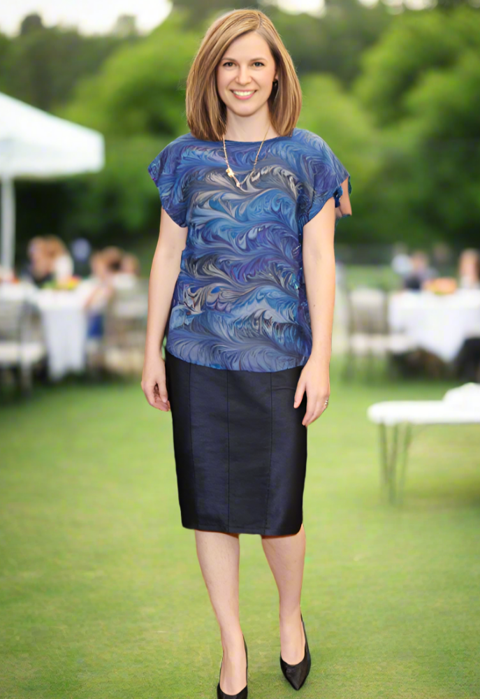 Woman in a marbled teal and gray top and black skirt standing in a formal setting with people in the background.