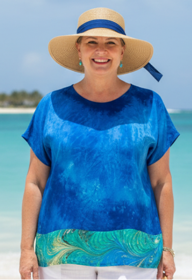 Woman in a blue hand dyed silk  top and white shorts standing on a beach with clear blue water and sky.