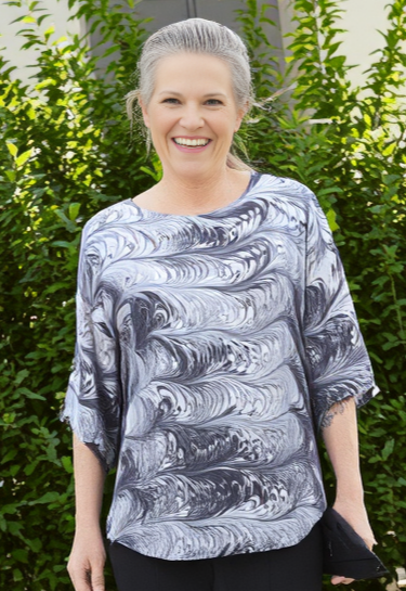 Woman wearing a marbled black and white silk top and black pants standing outdoors with greenery in the background