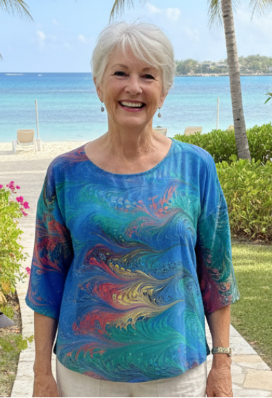 Woman wearing a half sleeve silk top marbled in blue, green, red, and yellow with the ocean in the background