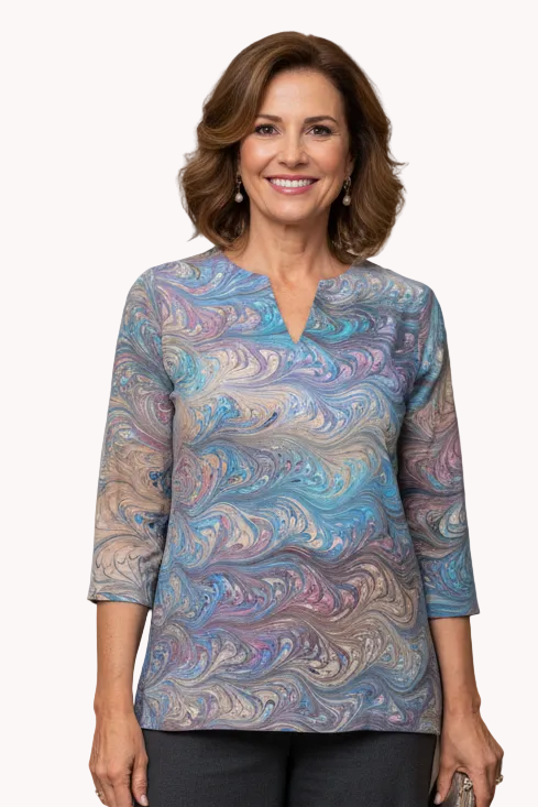 Woman in a marbled silk top and dark pants standing in an indoor setting with other people in the background.