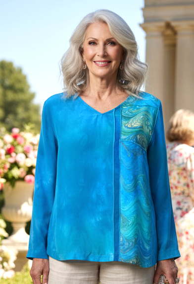 Woman in a hand dyed silk top with marbled inset and beige pants standing outdoors with floral decorations and people in the background.