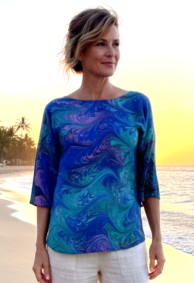 Woman standing on a beach wearing a blue and green marbled silk top and white pants.