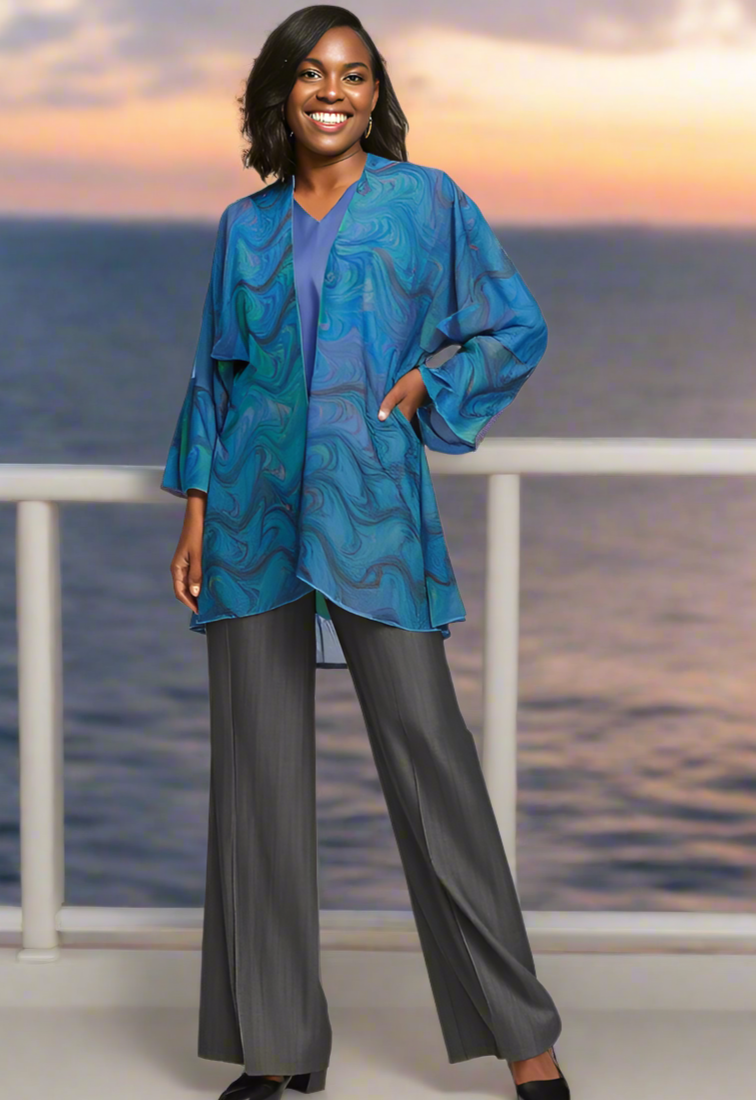 Woman wearing a blue marbled jacket and gray pants standing on a balcony with ocean view.