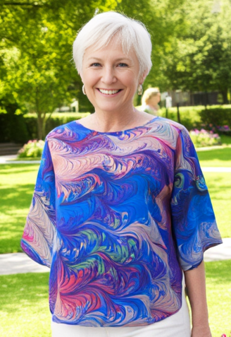 Woman in a colorful marbled silk top and white pants standing in a garden with flowers and greenery.