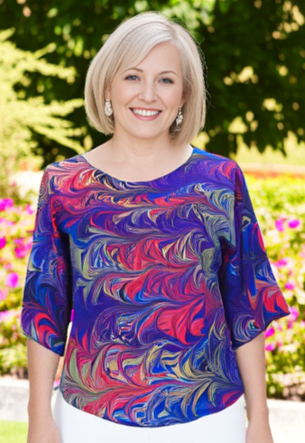 Woman wearing a colorful marbled silk top and white pants standing in a garden with flowers and greenery.