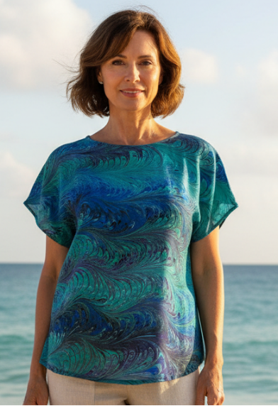 Woman standing on a beach wearing a blue marbled silk top and beige pants.