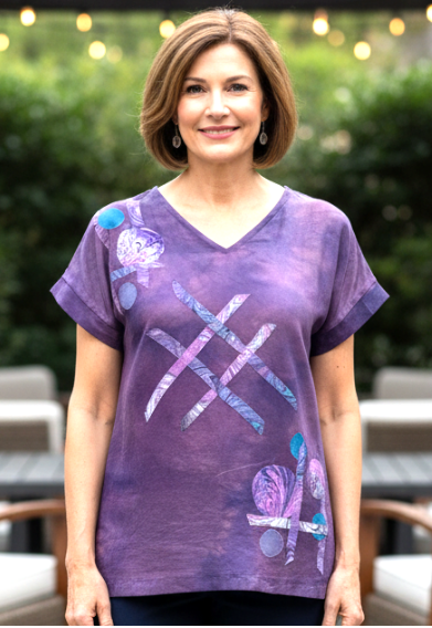 Woman wearing a hand dyed silk purple top with abstract designs and dark pants standing outdoors.