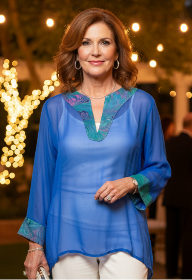 Woman in a sheer blue top with marbled trim and white pants standing outdoors with decorative lights in the background
