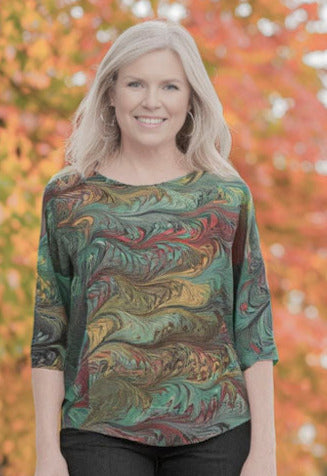 Woman wearing a marbled silk top and dark pants standing on a path with autumn trees in the background